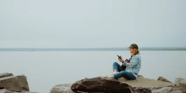 Pascale Picard plays guitar sitting on a rock by the river at the Quai de Saint-Michel-de-Bellechasse